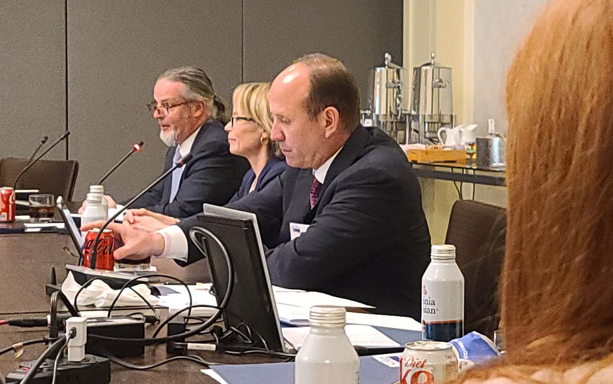 People sit at conference table in front of microphones and computer terminals