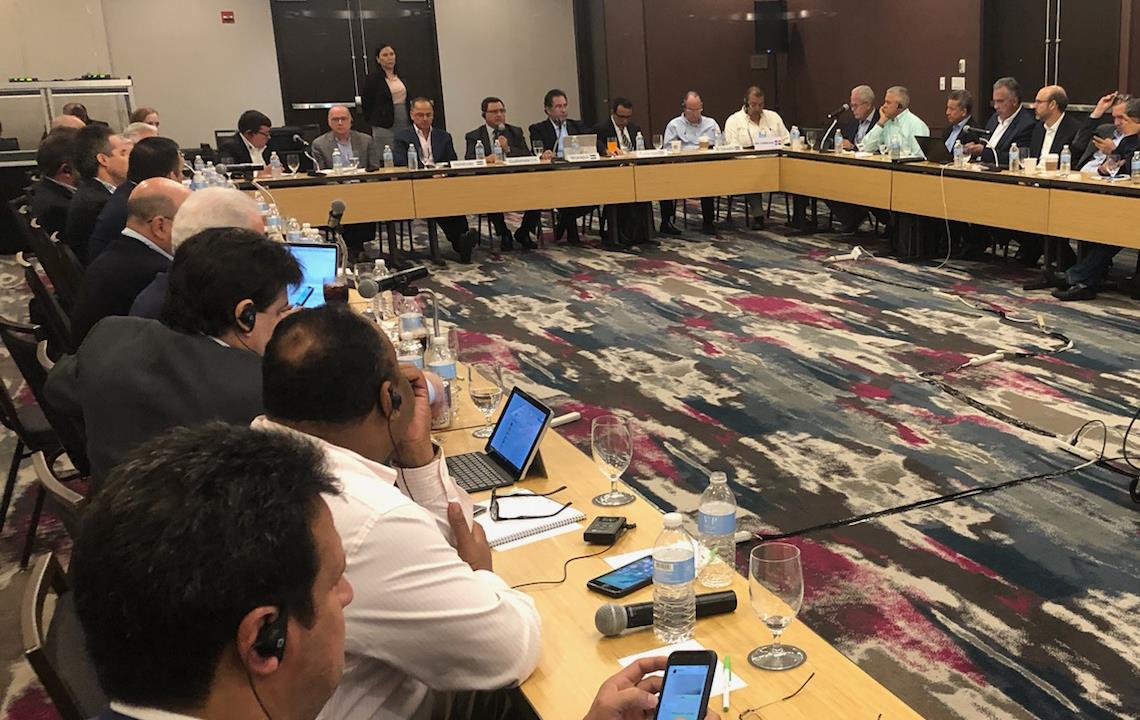 Large group gathered in conference room, seated at tables in U-shape, garish maroon, gray, white, and black rug dominates the photo