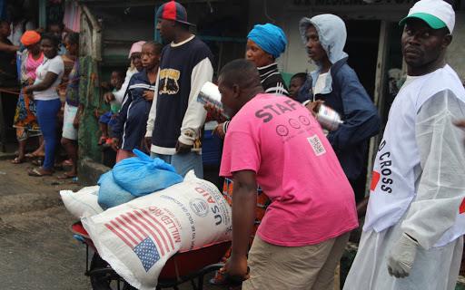 Man wearing pink t-shirt pushes wheelbarrow filled with bags of USAID milled rice in front of line of people