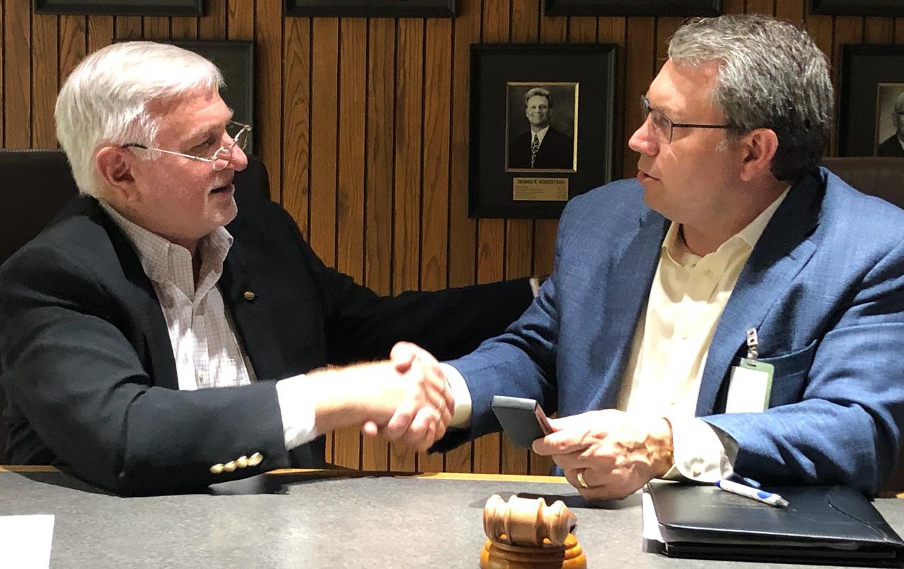 Two men wearing sport coats sit at table with gavel shaking hands, portrait photo gallery in background