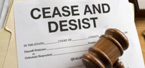 Wooden gavel covers piece of paper with text "Cease And Desist" atop stack of manila files
