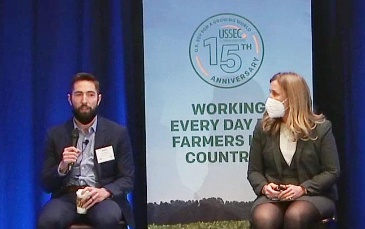 Man and woman sit in chairs on a stage in front of USSEC poster of soybeans