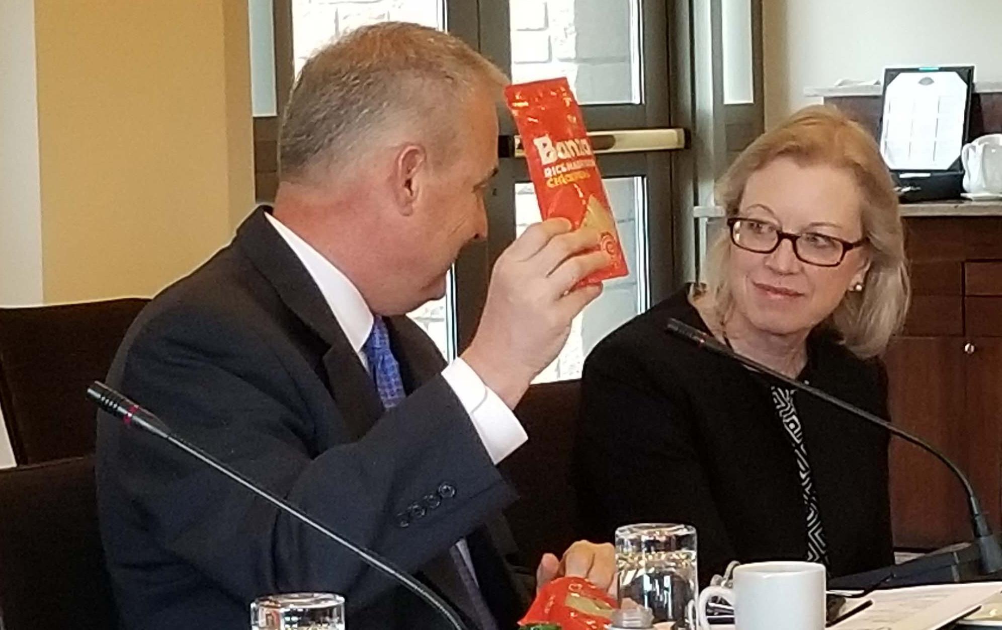 At a business meeting a man holds up a small, orange package while a woman wearing glasses smiles at him