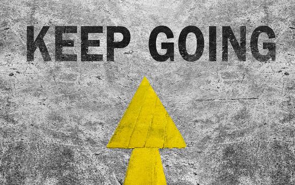 Black tennis shoes with white laces standing on yellow arrow pointed at text "Keep Going"