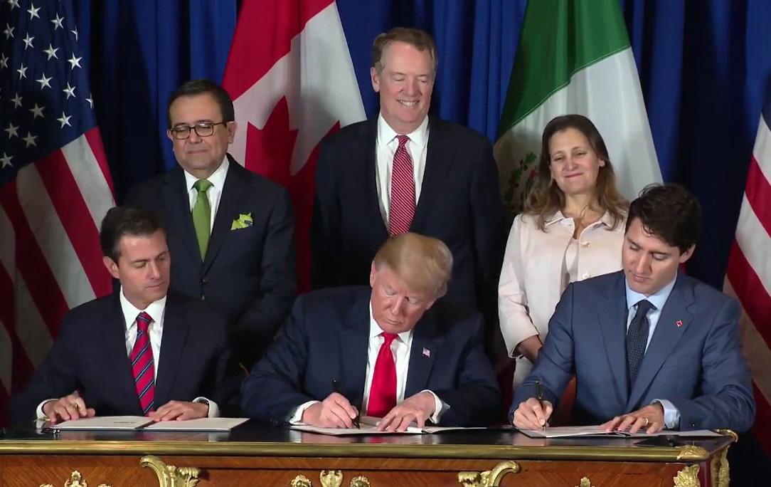 Three men wearing business suits sit at ornate desk signing documents, three people stand behind them looking on, country flags in background