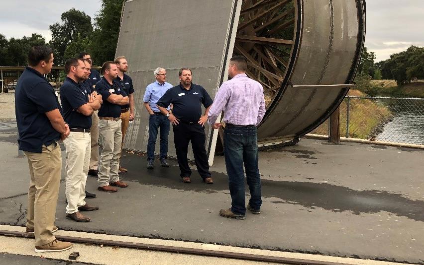 Group of men stand in front of enormous metal water wheel