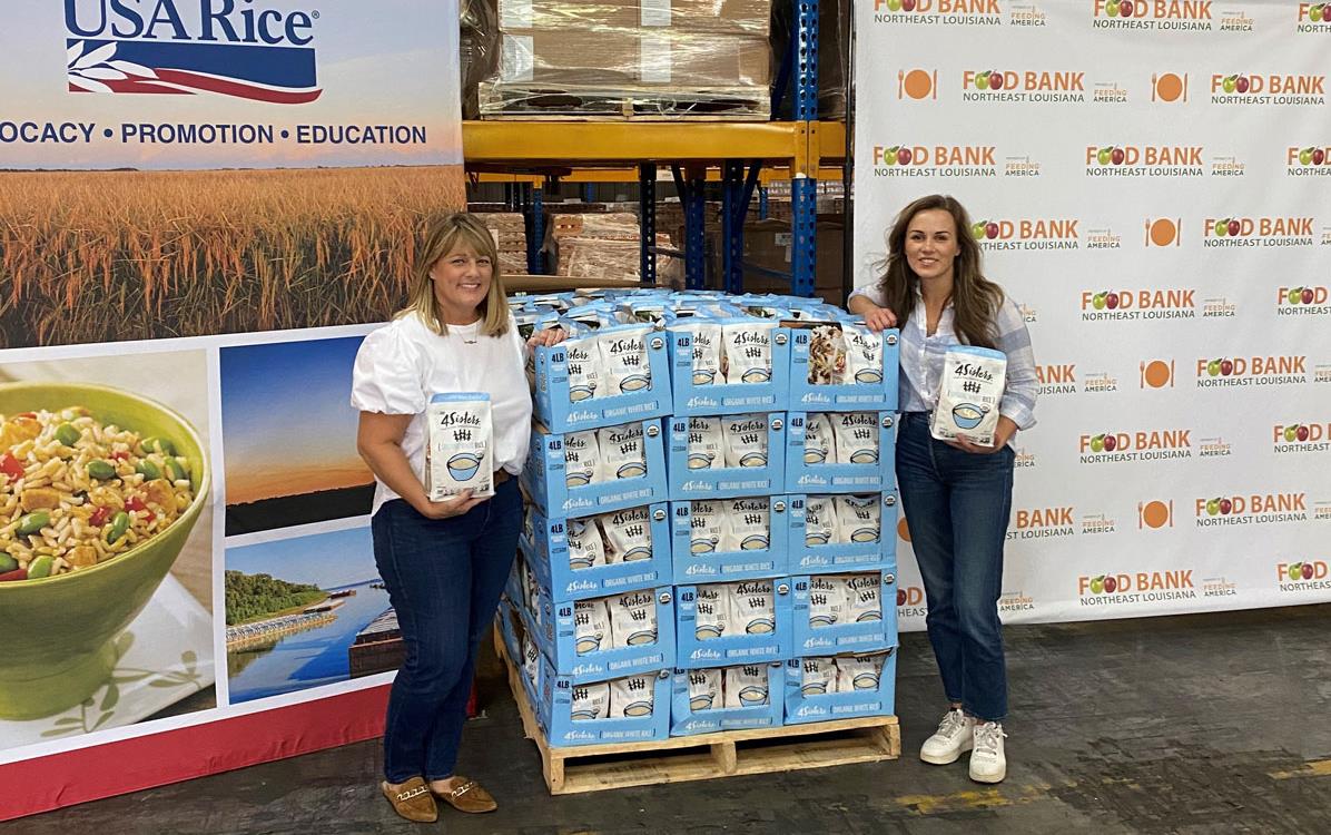 2022-4Sisters-NRM-Donation, two women, each holding a bag of rice, flank a pallet of rice bags