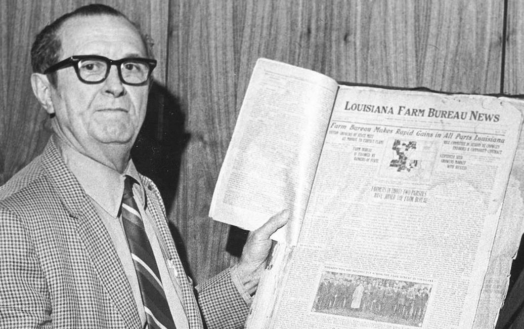 B/W photo of white man wearing horn-rimmed glasses holding LA Farm Bureau newspaper