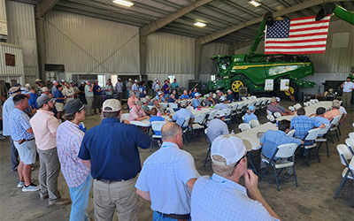 2026 Field Day at Richards Farm, crowd stands in front of combine & American flag hanging from ceiling