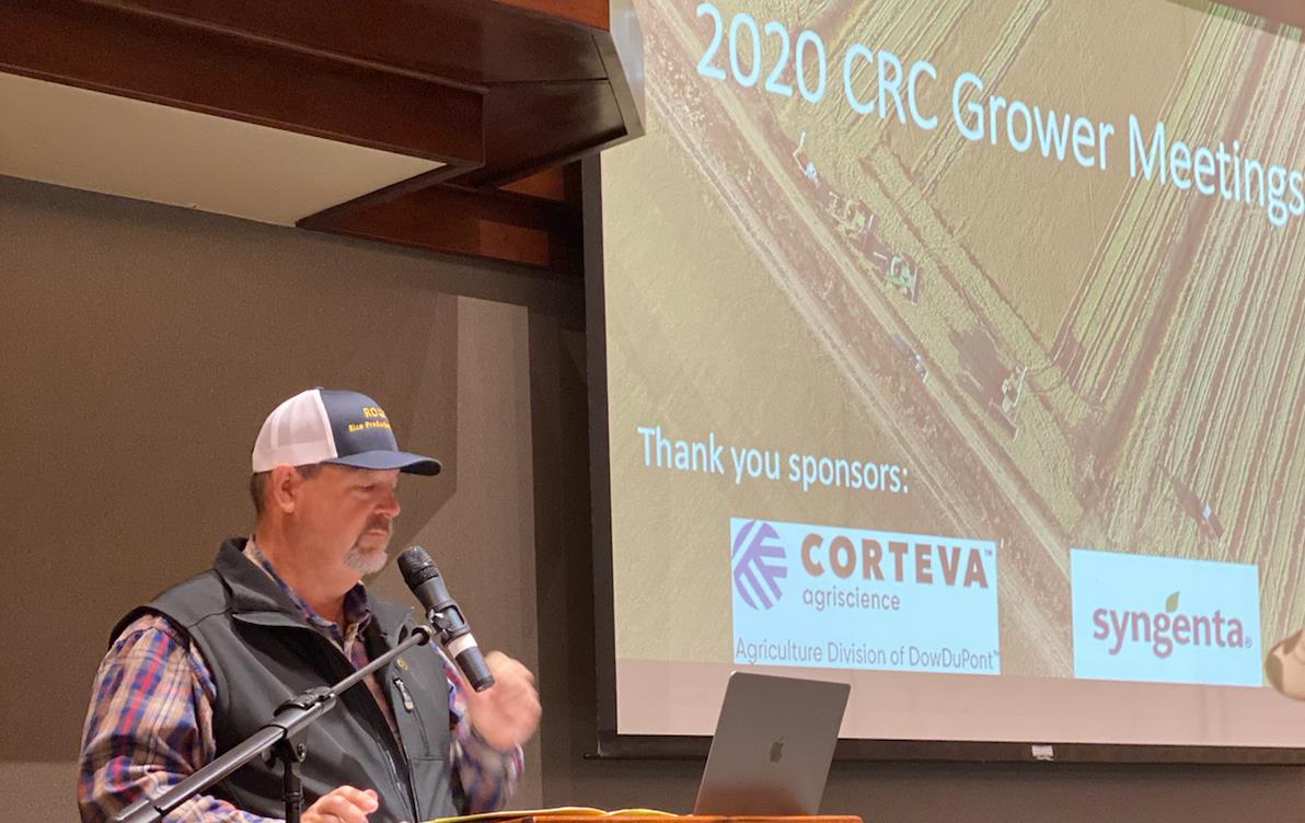 Casually dressed man wearing ballcap speaks from podium with slide presentation in background