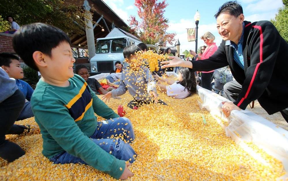Children play in a large container filled with yellow corn, woman throws corn in the air