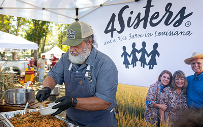 2025 Rice Bowl Game, 4Sisters Tailgate Booth, man wearing apron and ballcap spoons out rice dish