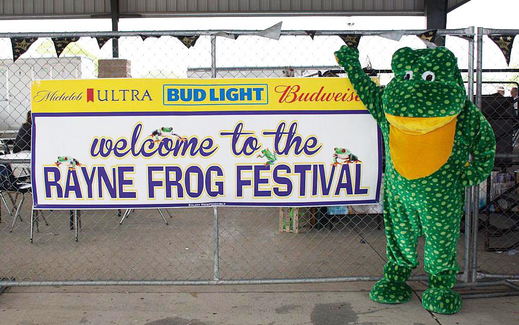 Green frog character stands next to "Welcome-to-the-Rayne-Frog-Festival" sign