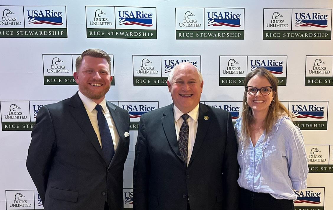 Duck-Stamp-Reception-J. Cruce, Rep Ron Estes & DUs Julia Peebles stand in front of USAR & DU step and repeat