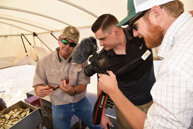 Cameraman films man in ball cap and sunglasses holding two crawfish while another man wearing a black shirt looks on