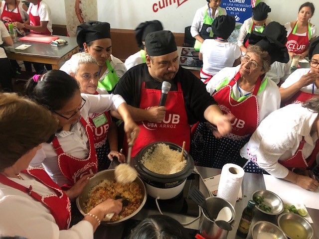 Chef Javier Rodriguez in the middle of a crowd of women wearing red Club Arroz aprons, preparing dish using US rice