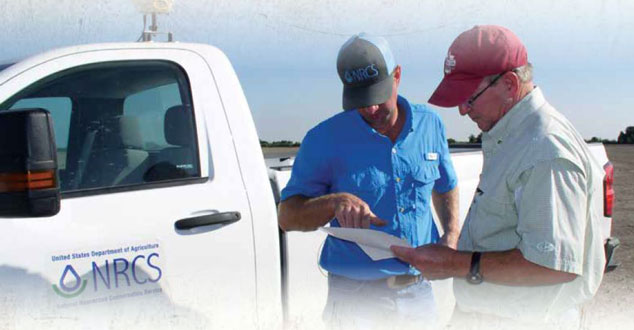 Two men standing near a white pick-up truck with the NRCS logo on the door, one holds a piece of paper and the other is pointing to it