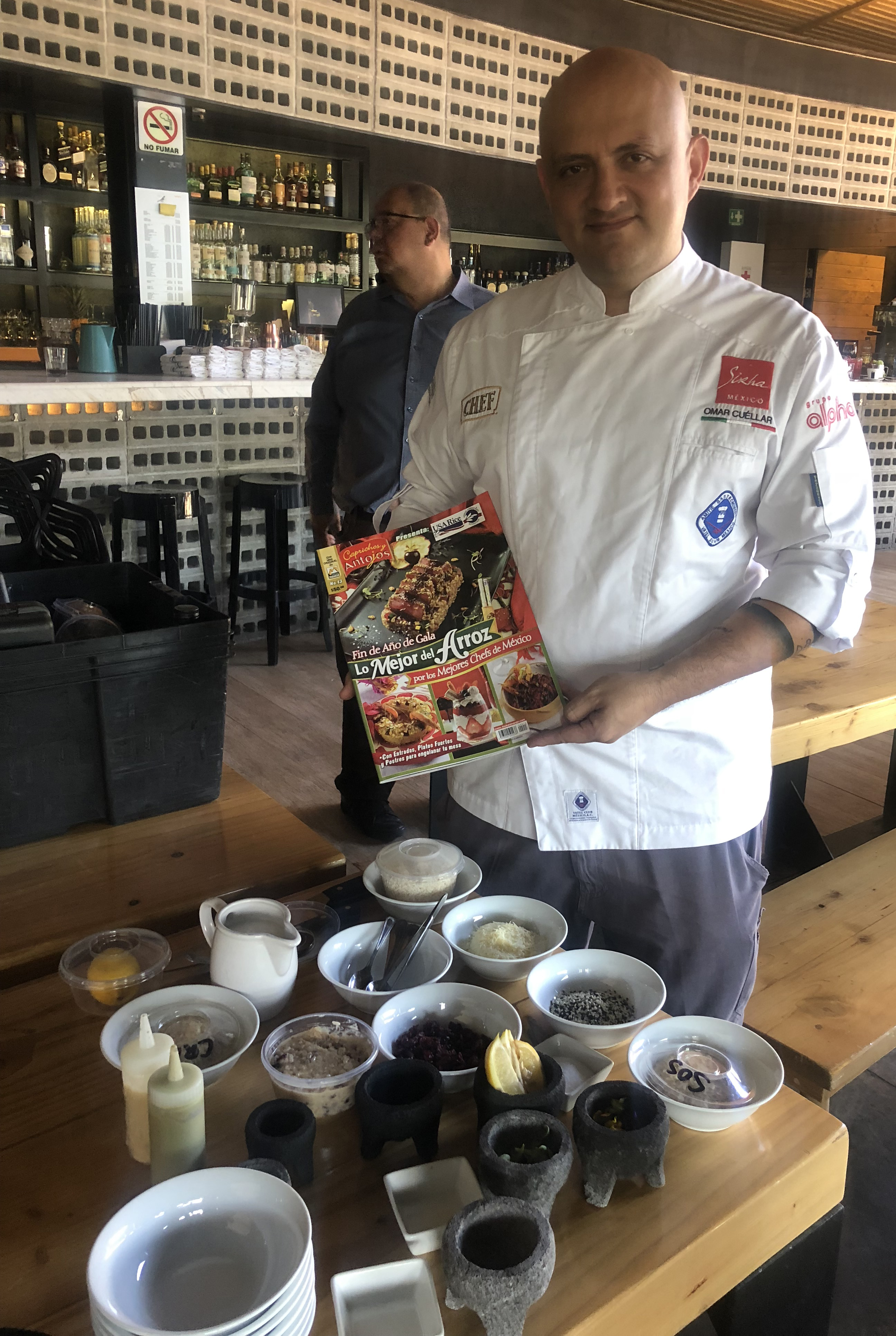 Chef stands in front of table full of ingredients, holding cookbook