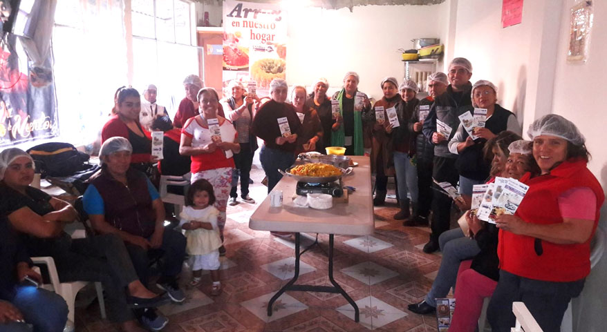 Group shot of seminar participants in Mexican soup kitchen, people in hairnets holding rice pamphlets surround food table